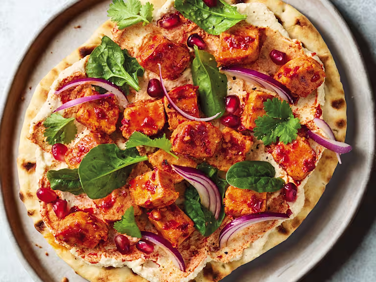 Harissa-spiced vegan Quorn pieces on a flatbread with hummus, pomegranate and coriander on a white plate with a blue background.