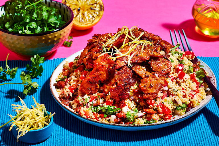 A couscous salad with red onion, red pepper, and tomato topped with Quorn Makes Amazing Peri Peri Strips on a turquoise placemat against a fuchsia background.