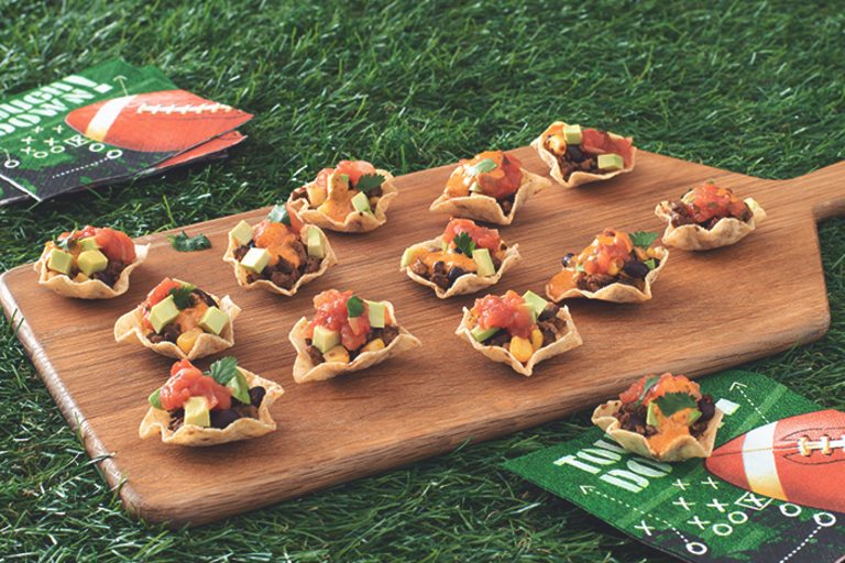 Mini Tortilla Chip Bowls Filled with Quorn Grounds, black beans, salsa, queso, avocado, and cilantro arranged on a board with game day napkins placed around it.