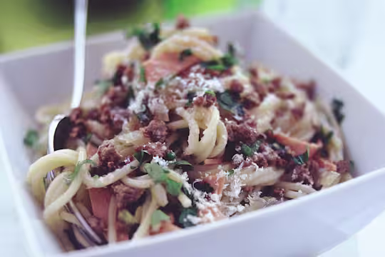 Close up shot of Quorn Mince and spaghetti in a square white bowl garnished with parsley and parmesan cheese.