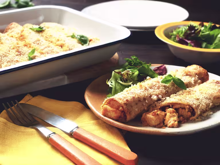 Two baked pancakes filled with Quorn Pieces and vegetables topped with breadcrumbs and cheese plated with a side salad and the full baking dish in the background.