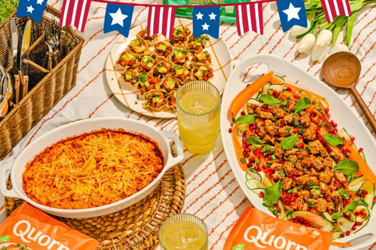 A table spread of Quorn Vegetarian 4th of July recipes on a stripy tablecloth next to a wicker basket and glasses of orange juice.