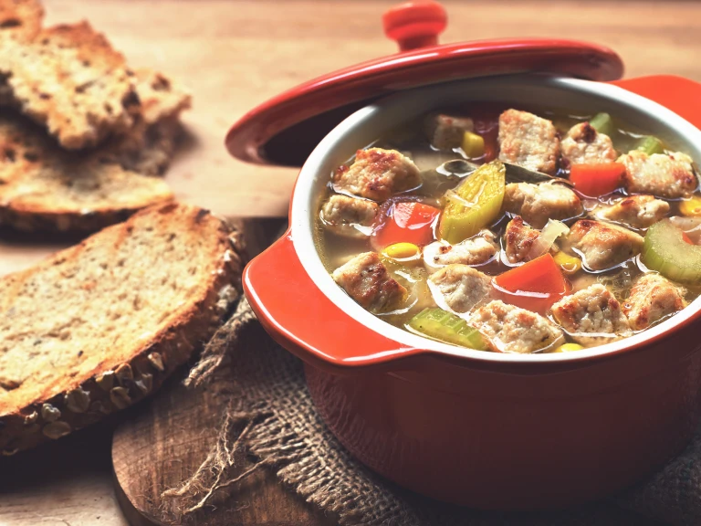 A vegetable soup with carrots, celery, sweetcorn, and Quorn Pieces in a red Dutch oven with toasted wholegrain bread on the side.