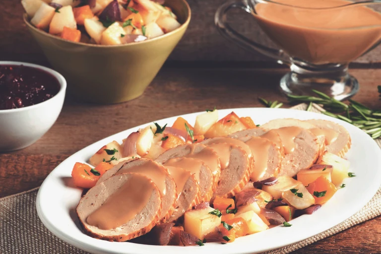 Sliced Quorn Meatless Vegetarian Turkey Roast topped with gravy with roasted root vegetables on the side on an oblong-shaped white plate.
