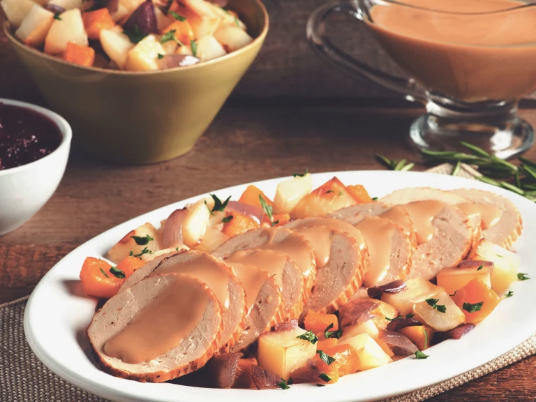 Sliced Quorn Meatless Vegetarian Turkey Roast topped with gravy with roasted root vegetables on the side on an oblong-shaped white plate.