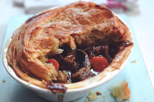 A golden, flaky vegetarian steak and ale pie filled with rich gravy, mushrooms, carrots, and Quorn Steak Strips, served in a white dish on a light blue surface