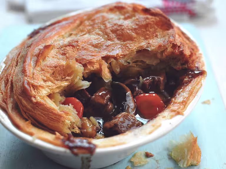 A golden, flaky vegetarian steak and ale pie filled with rich gravy, mushrooms, carrots, and Quorn Steak Strips, served in a white dish on a light blue surface