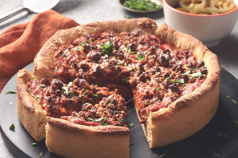 Chicago-Style Deep Dish Pizza topped with Quorn Meatless Grounds and cheese, on a black dish alongside an orange napkin and bowls of toppings like jalapenos.