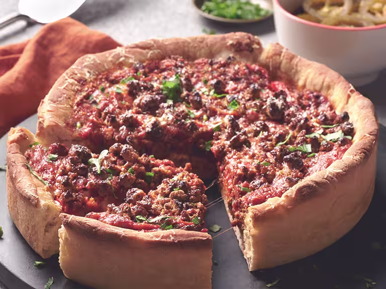 Chicago-Style Deep Dish Pizza topped with Quorn Meatless Grounds and cheese, on a black dish alongside an orange napkin and bowls of toppings like jalapenos.