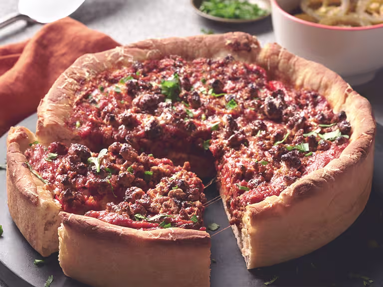 Chicago-Style Deep Dish Pizza topped with Quorn Meatless Grounds and cheese, on a black dish alongside an orange napkin and bowls of toppings like jalapenos.