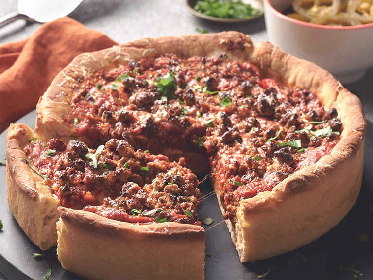 Chicago-Style Deep Dish Pizza topped with Quorn Meatless Grounds and cheese, on a black dish alongside an orange napkin and bowls of toppings like jalapenos.