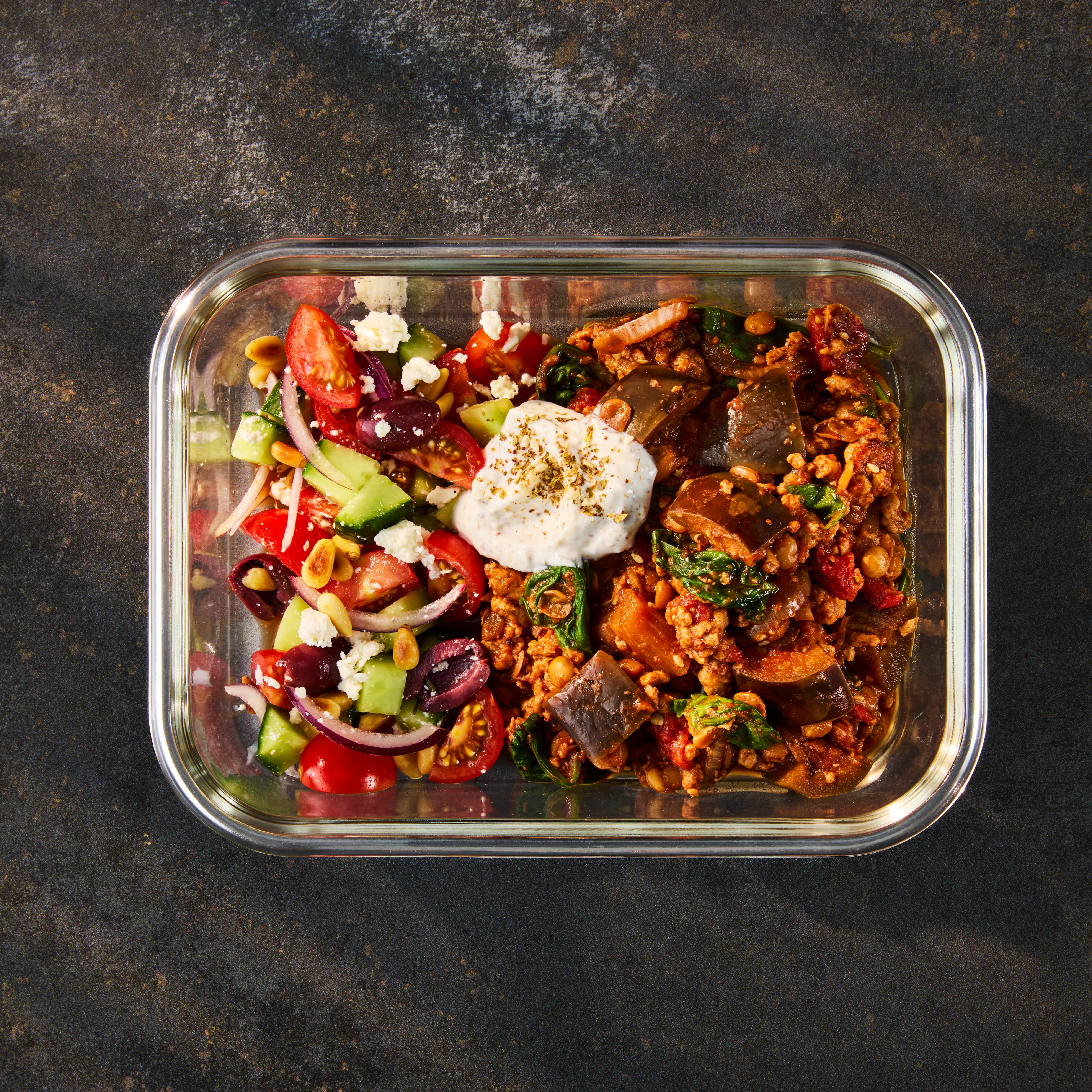 A glass meal prep container holding Quorn Mince Moussaka alongside a fresh Greek Salad and herbed yogurt.