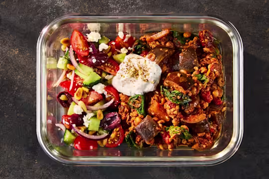 A glass meal prep container holding Quorn Mince Moussaka alongside a fresh Greek Salad and herbed yogurt.