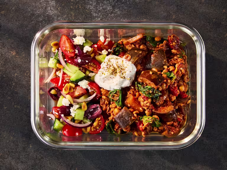 A glass meal prep container holding Quorn Mince Moussaka alongside a fresh Greek Salad and herbed yogurt.