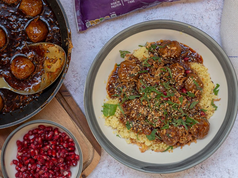 Quorn Swedish Style Balls in a bowl on a bed of cous cous topped with fresh herbs. The meat-free meatballs are also in a pan next to the bowl along with a small dish of pomegranate. 