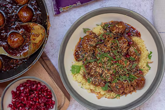 Quorn Swedish Style Balls in a bowl on a bed of cous cous topped with fresh herbs. The meat-free meatballs are also in a pan next to the bowl along with a small dish of pomegranate.
