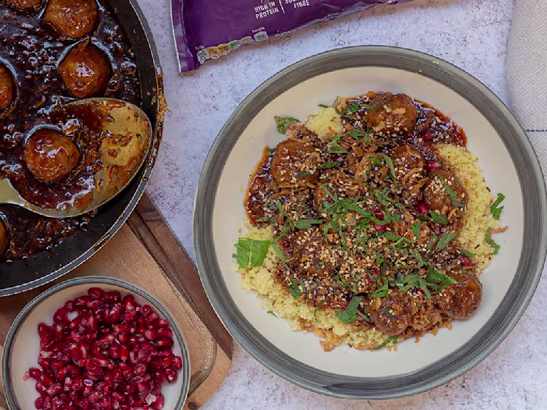 Quorn Swedish Style Balls in a bowl on a bed of cous cous topped with fresh herbs. The meat-free meatballs are also in a pan next to the bowl along with a small dish of pomegranate.