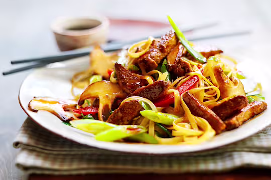A serving of vegetarian teriyaki noodles made with Quorn Vegetarian Steak Strips, mixed vegetables, and teriyaki sauce on a white plate with chopsticks on the side.