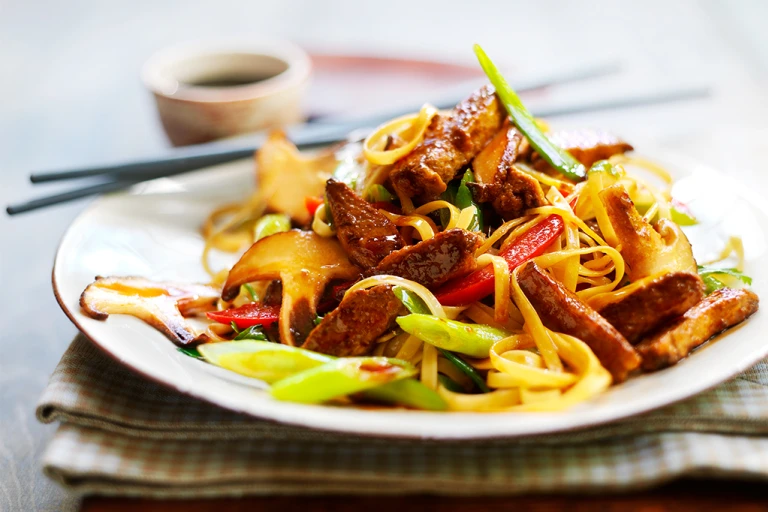 A serving of vegetarian teriyaki noodles made with Quorn Vegetarian Steak Strips, mixed vegetables, and teriyaki sauce on a white plate with chopsticks on the side.