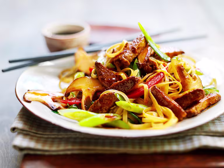 A serving of vegetarian teriyaki noodles made with Quorn Vegetarian Steak Strips, mixed vegetables, and teriyaki sauce on a white plate with chopsticks on the side.