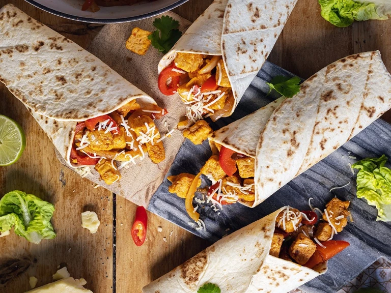 Four vegetarian fajita wraps made with Quorn Pieces, peppers, and onions on a board surrounded by the fillings and veggies.