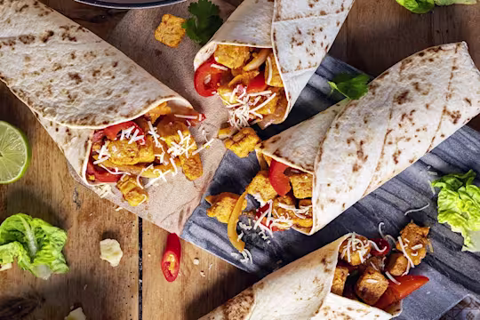Four vegetarian fajita wraps made with Quorn Pieces, peppers, and onions on a board surrounded by the fillings and veggies.
