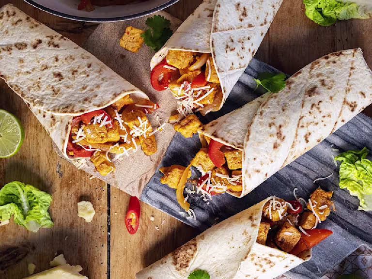 Four vegetarian fajita wraps made with Quorn Pieces, peppers, and onions on a board surrounded by the fillings and veggies.