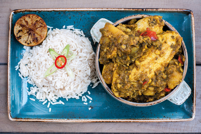 A mini cocotte of Trini curry chicken made with Quorn Vegan Pieces served with a side of rice and half of a charred citrus on a blue tray.