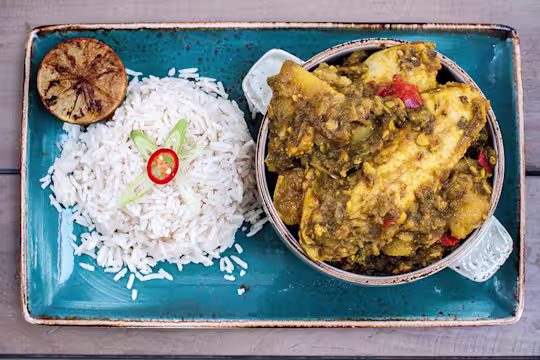 A mini cocotte of Trini curry chicken made with Quorn Vegetarian Fillets served with a side of rice and half of a charred citrus on a blue tray.
