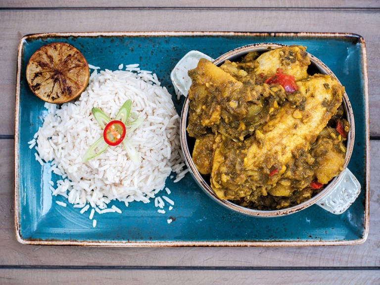 A mini cocotte of Trini curry chicken made with Quorn Vegetarian Fillets served with a side of rice and half of a charred citrus on a blue tray.