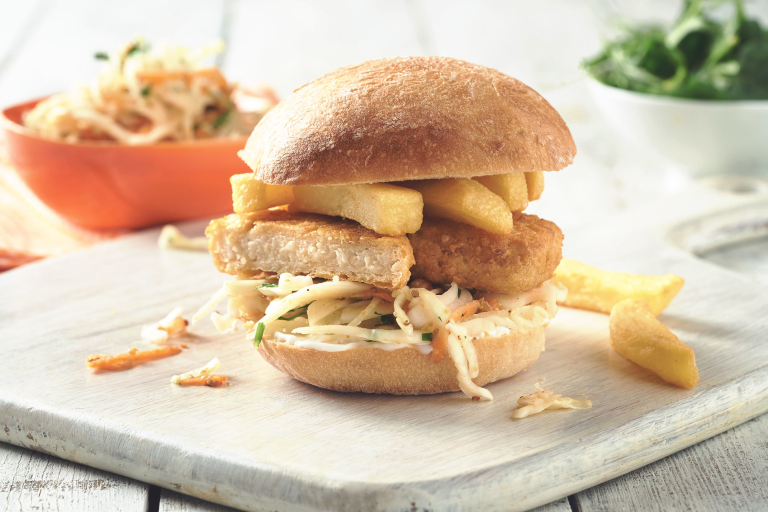 Quorn Vegan Fishless Fingers sandwiched in a bun with coleslaw and chips served on a white board with a few scattered chips.