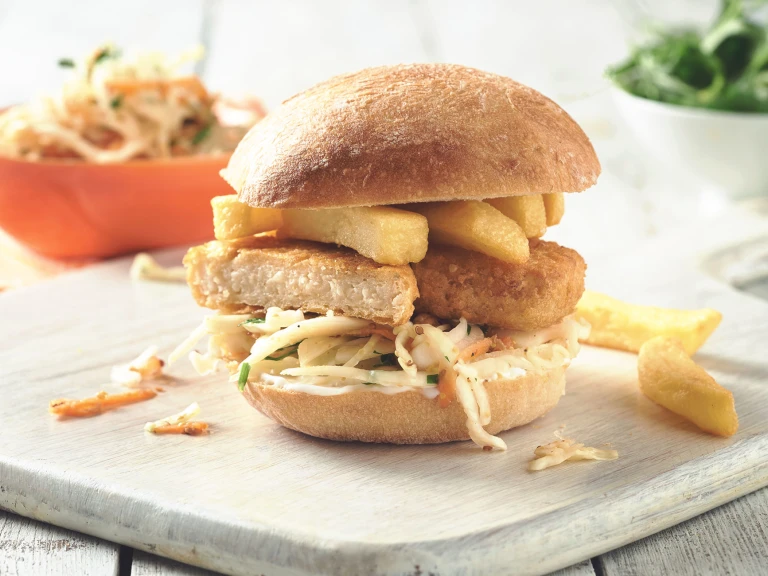 Quorn Vegan Fishless Fingers sandwiched in a bun with coleslaw and chips served on a white board with a few scattered chips.