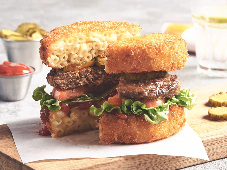 A veggie burger topped with lettuce, tomatoes, pickles, and ketchup with two breaded patties of macaroni and cheese as a bun, served on a plank.