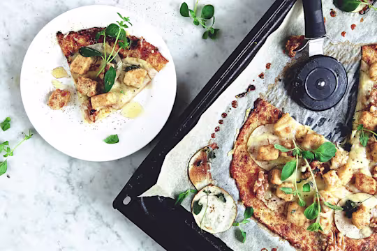 A flatbread topped with cheese, pears, walnuts, and Quorn Pieces on a baking sheet with one slice served.