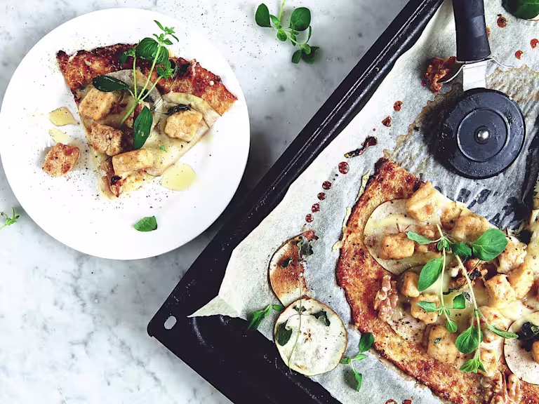 A flatbread topped with cheese, pears, walnuts, and Quorn Pieces on a baking sheet with one slice served.