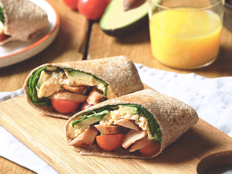 Quorn Vegetarian Breakfast Burrito, made with Quorn Sausages, tomato, spinach and served in a wholemeal wrap on a board with a glass of orange juice.