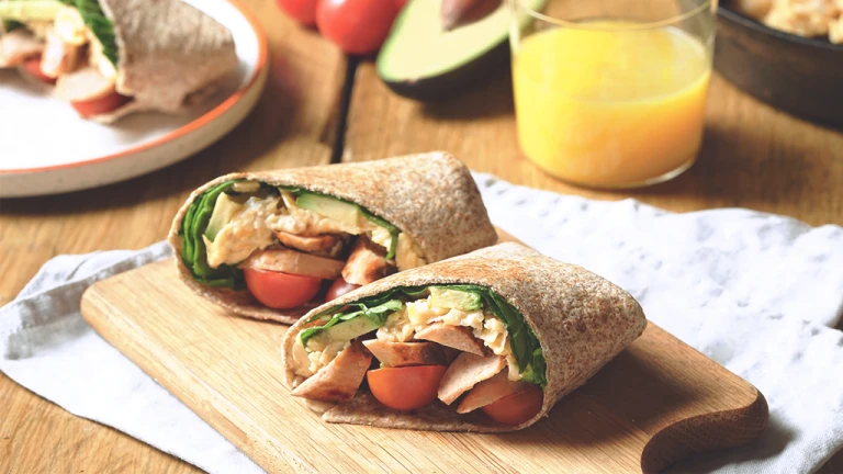 Quorn Vegetarian Breakfast Burrito, made with Quorn Sausages, tomato, spinach and served in a wholemeal wrap on a board with a glass of orange juice.