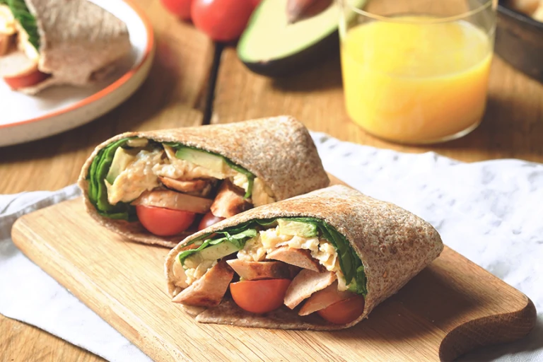 Quorn Vegetarian Breakfast Burrito, made with Quorn Sausages, tomato, spinach and served in a wholemeal wrap on a board with a glass of orange juice.