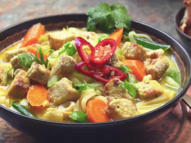 A bowl of Thai Noodle Soup with Quorn Vegan Pieces topped with sliced red chilli and sat beside a bowl of crunchy bread.