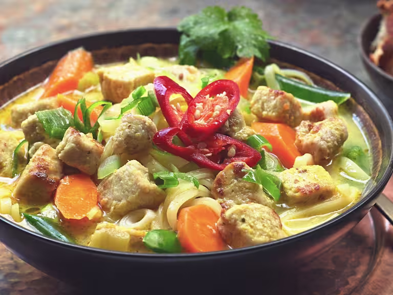 A bowl of Thai Noodle Soup with Quorn Vegan Pieces topped with sliced red chilli and sat beside a bowl of crunchy bread.