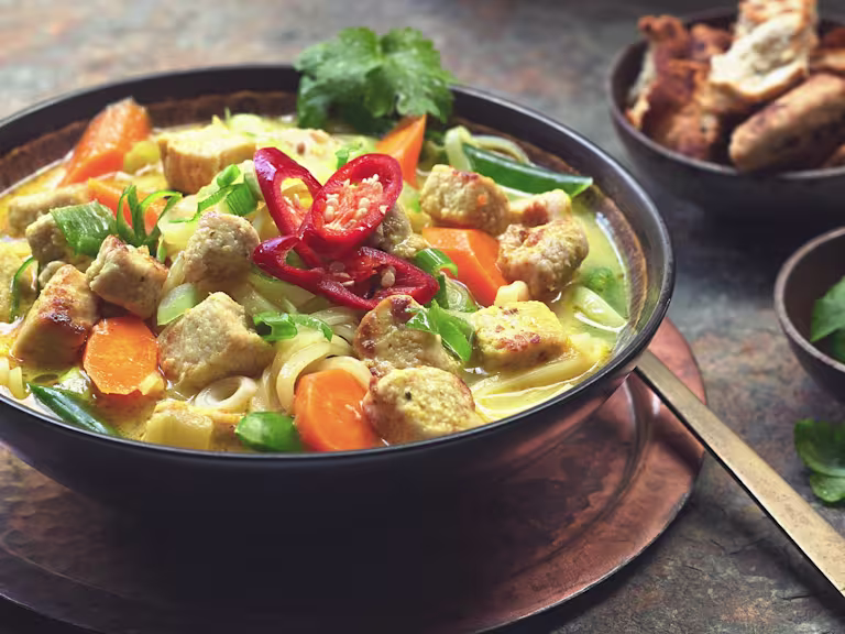 A bowl of Thai Noodle Soup with Quorn Vegan Pieces topped with sliced red chilli and sat beside a bowl of crunchy bread.