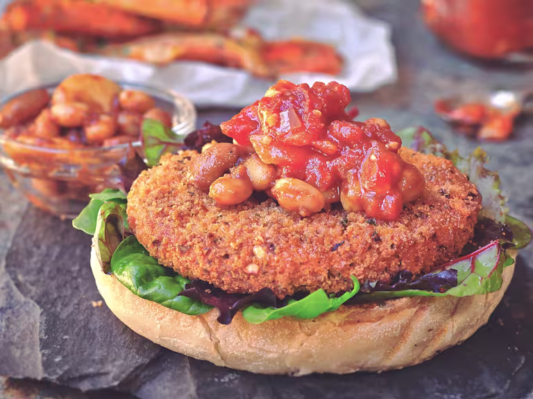 Quorn Vegan Hot and Spicy Burger layered on top of salad and half a bun topped with bbq beans with a pot of beans on the side
