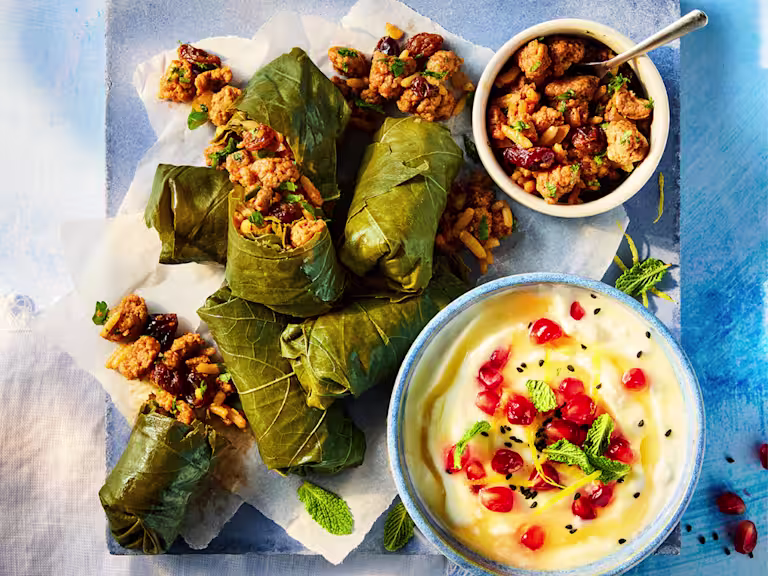 Vegetarian dolmades served on white paper with a feta dip topped with pomegranate seeds and a bowl of filling to the side.