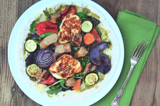An open wrap topped with pesto, red onions, courgette, squash, red peppers, grilled halloumi, and Quorn Sausages on a white plate.