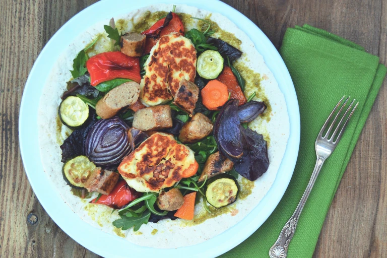 An open wrap topped with pesto, red onions, courgette, squash, red peppers, grilled halloumi, and Quorn Sausages on a white plate.