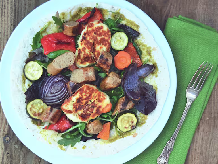 An open wrap topped with pesto, red onions, courgette, squash, red peppers, grilled halloumi, and Quorn Sausages on a white plate.
