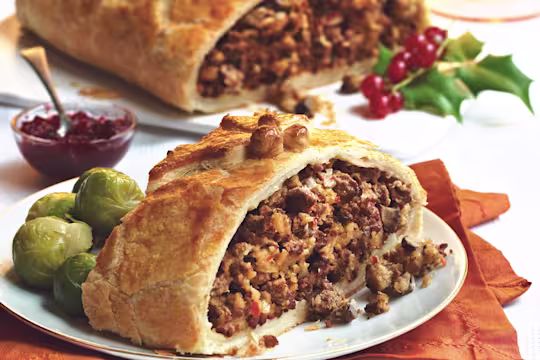A slice of puff pastry filled with Quorn Grounds and mushrooms with Brussels sprouts on the side, and a chutney and the larger Wellington from which the slice has been taken in the background.