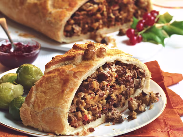 A slice of puff pastry filled with Quorn Grounds and mushrooms with Brussels sprouts on the side, and a chutney and the larger Wellington from which the slice has been taken in the background.