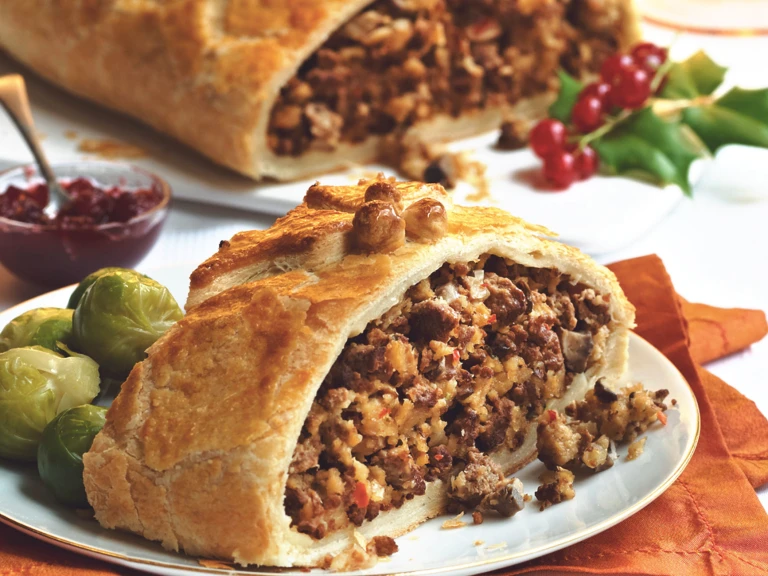 A slice of puff pastry filled with Quorn Grounds and mushrooms with Brussels sprouts on the side, and a chutney and the larger Wellington from which the slice has been taken in the background.