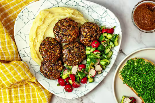 Five vegetarian Frikadellen meatballs on a bed of mashed potatoes with chopped vegetables served on a white plate with a side dip and bread.